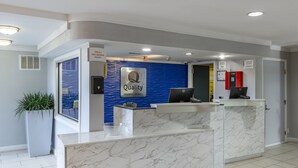 Zona de recepción de hotel con mostrador de mármol, pared azul con un letrero de 'Quality by Choice Hotels' y una computadora sobre el mostrador.