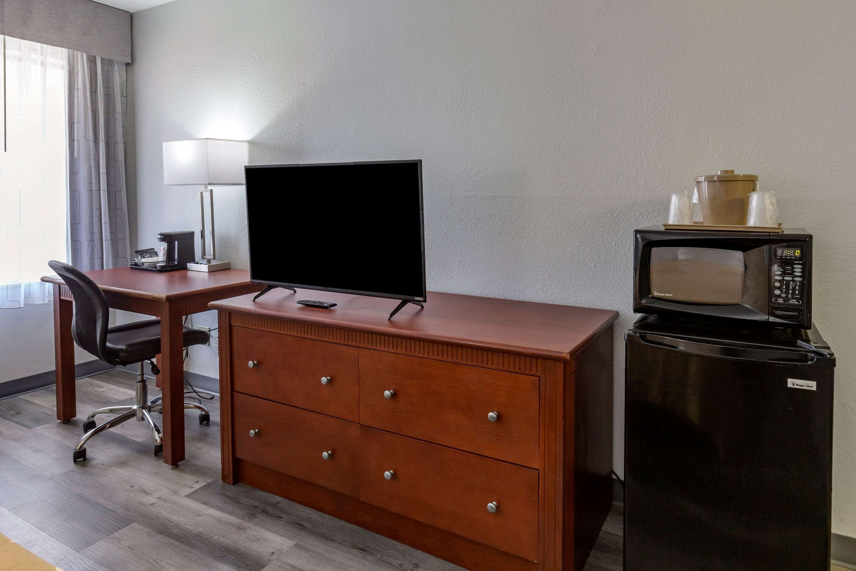 A hotel room with a wooden desk, a black TV, a microwave, a refrigerator, a chair, and a lamp.