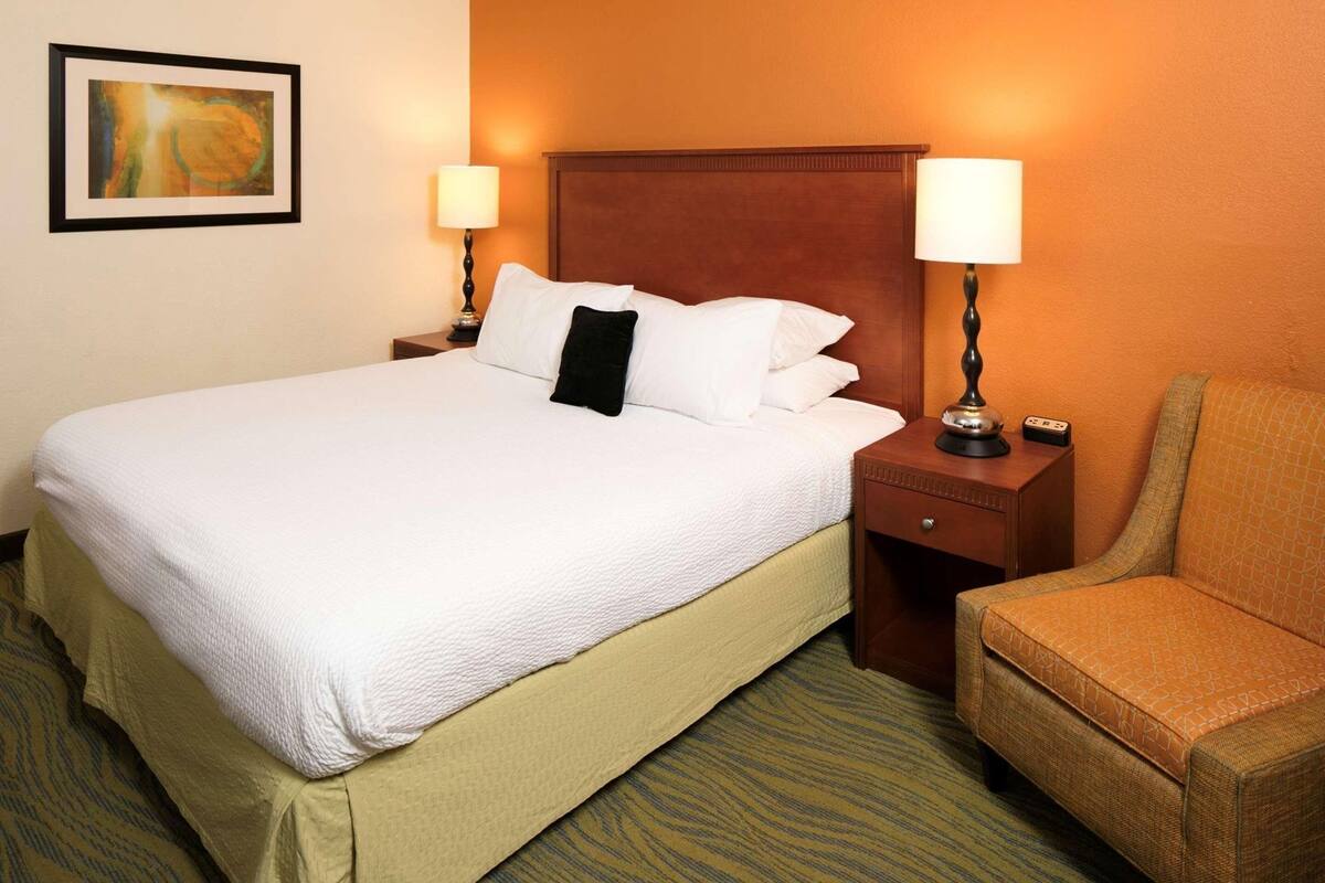 A hotel room with a bed, two bedside lamps, a chair, and a painting on the wall.