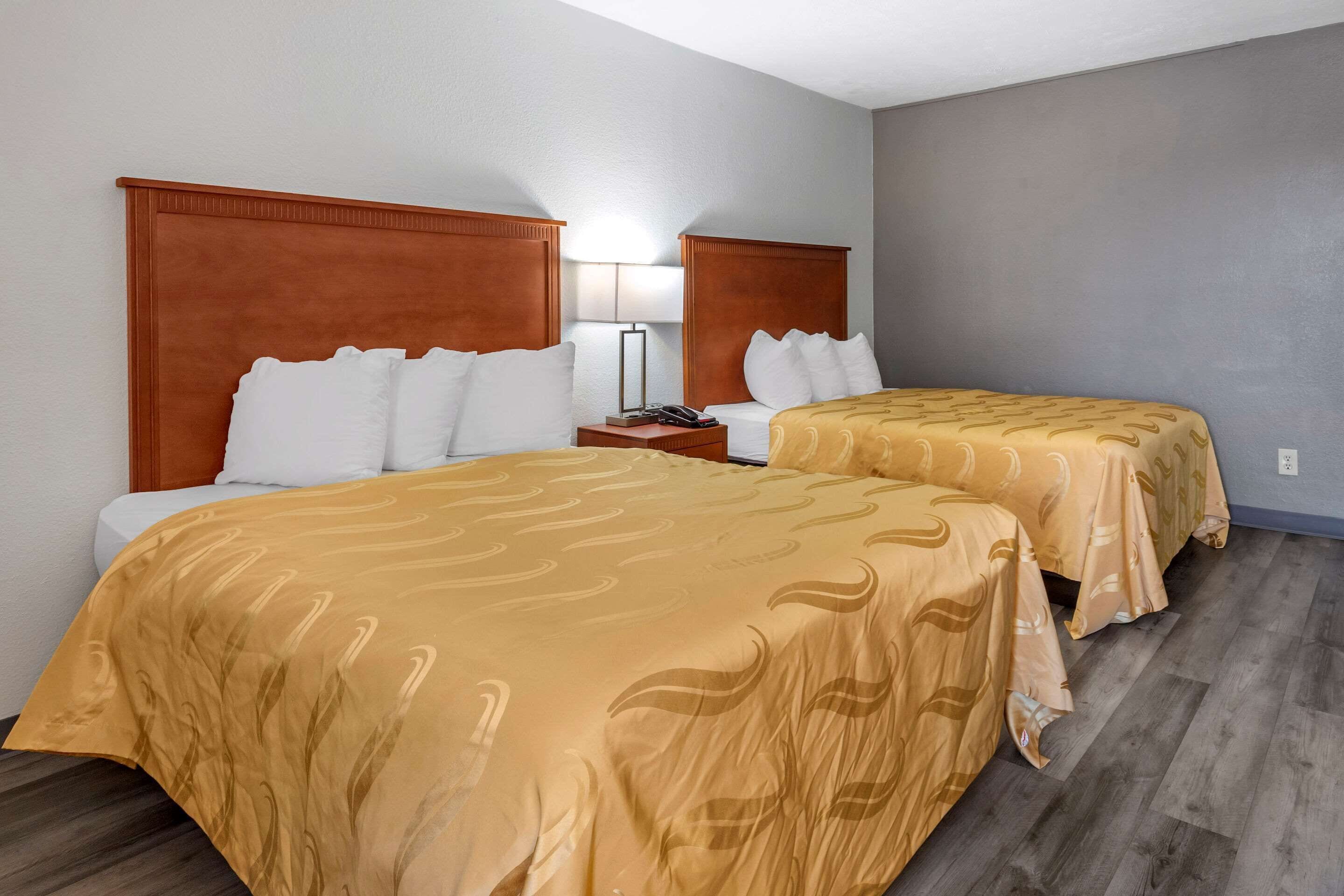 A hotel room with two beds, each with a wooden headboard and white bedding. There is a nightstand with a lamp and a phone between the beds. The room has a gray wall and a wooden floor.