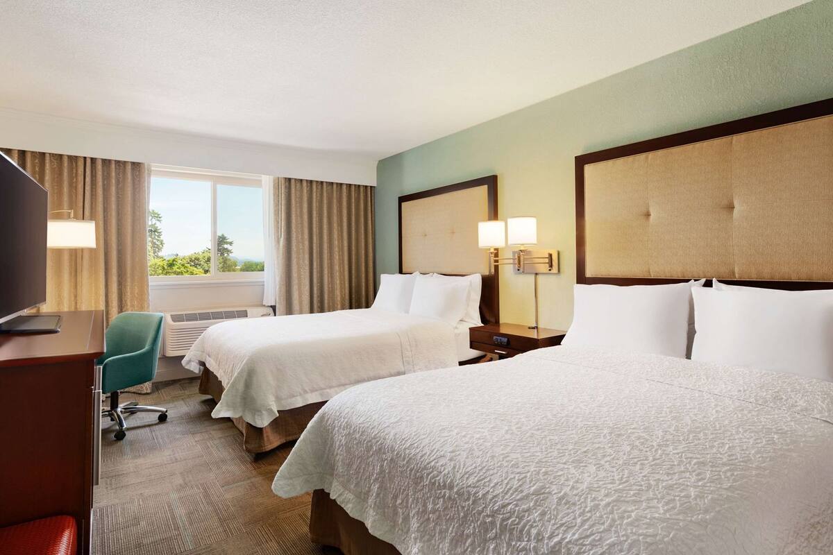 A hotel room with two beds, a desk, a chair, a TV, and a window with curtains.