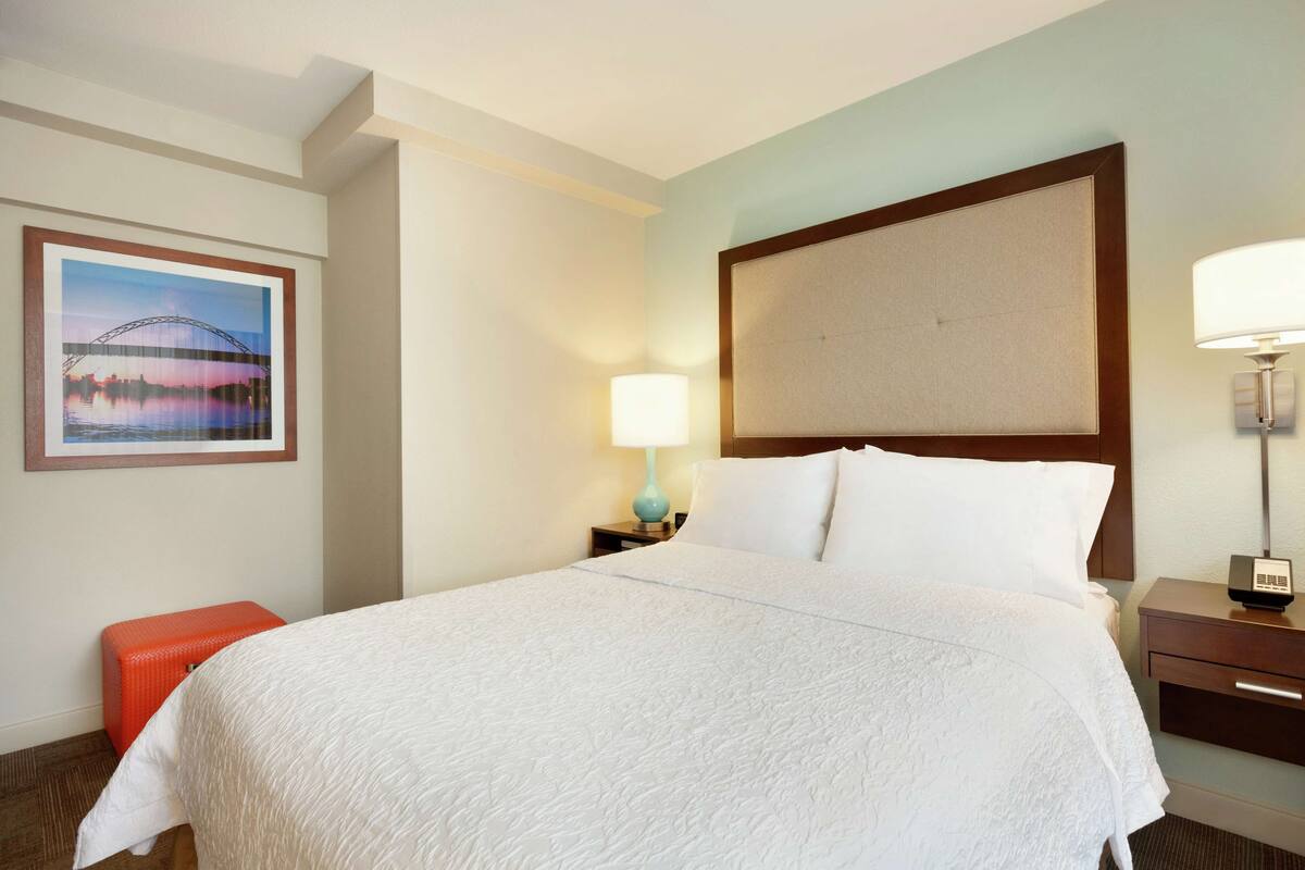 A hotel room with a large bed, a nightstand with a lamp, a framed picture on the wall, and a small orange stool.