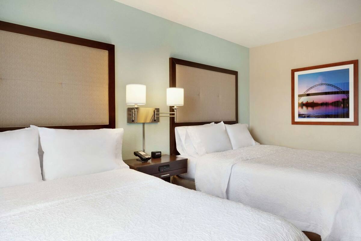A hotel room with two beds, a nightstand with a phone and lamp, and a framed picture on the wall.