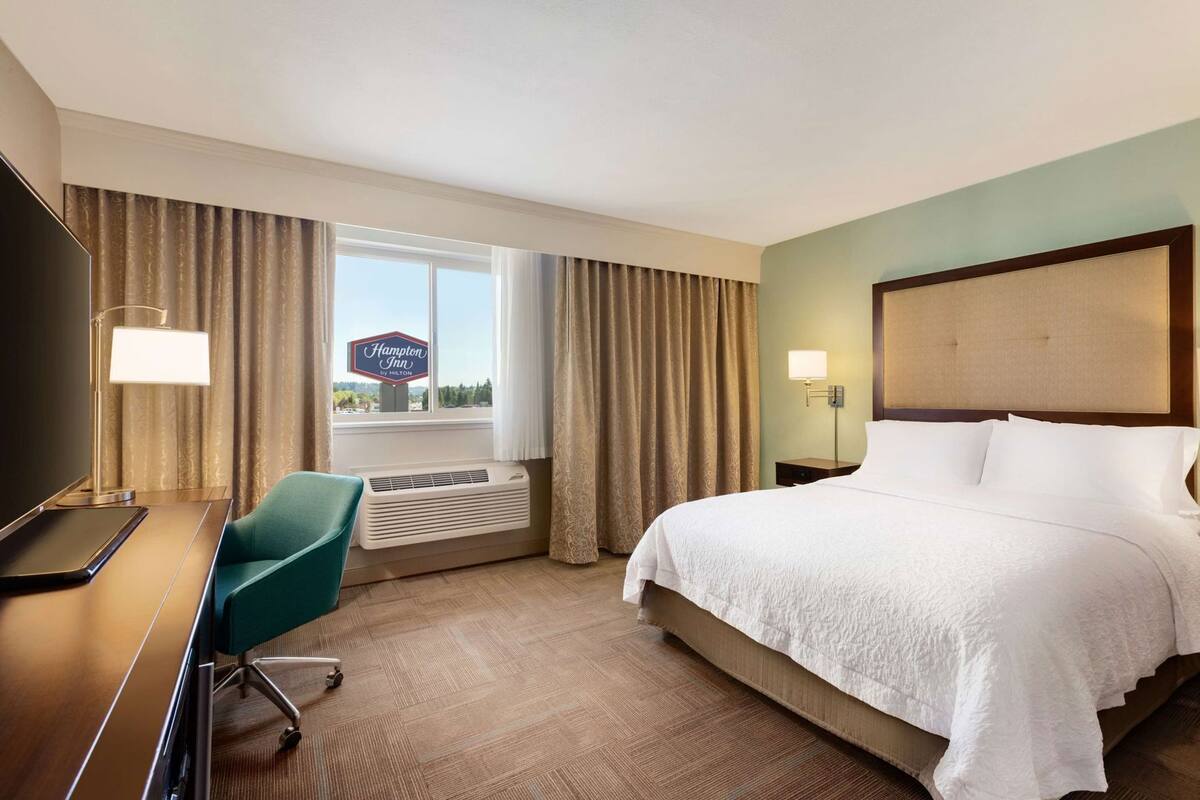 A hotel room with a bed, a desk, a chair, a TV, a sign for Hampton Inn by Hilton, and a window with curtains.