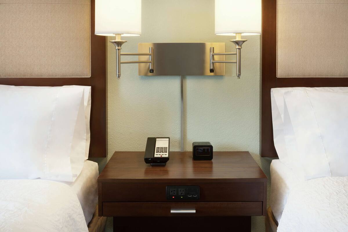 A hotel room with a wooden nightstand, two bedside lamps, a digital clock, and a phone.