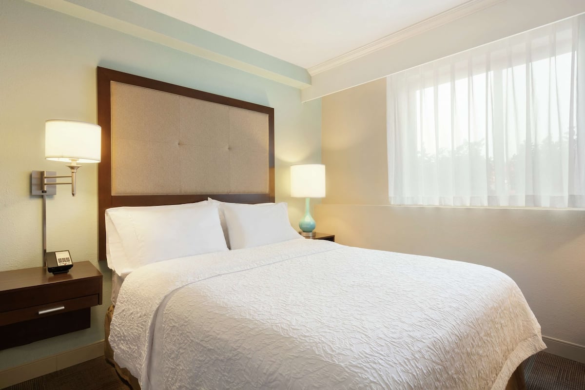 A hotel room with a large bed, two bedside tables, and a window with sheer curtains.