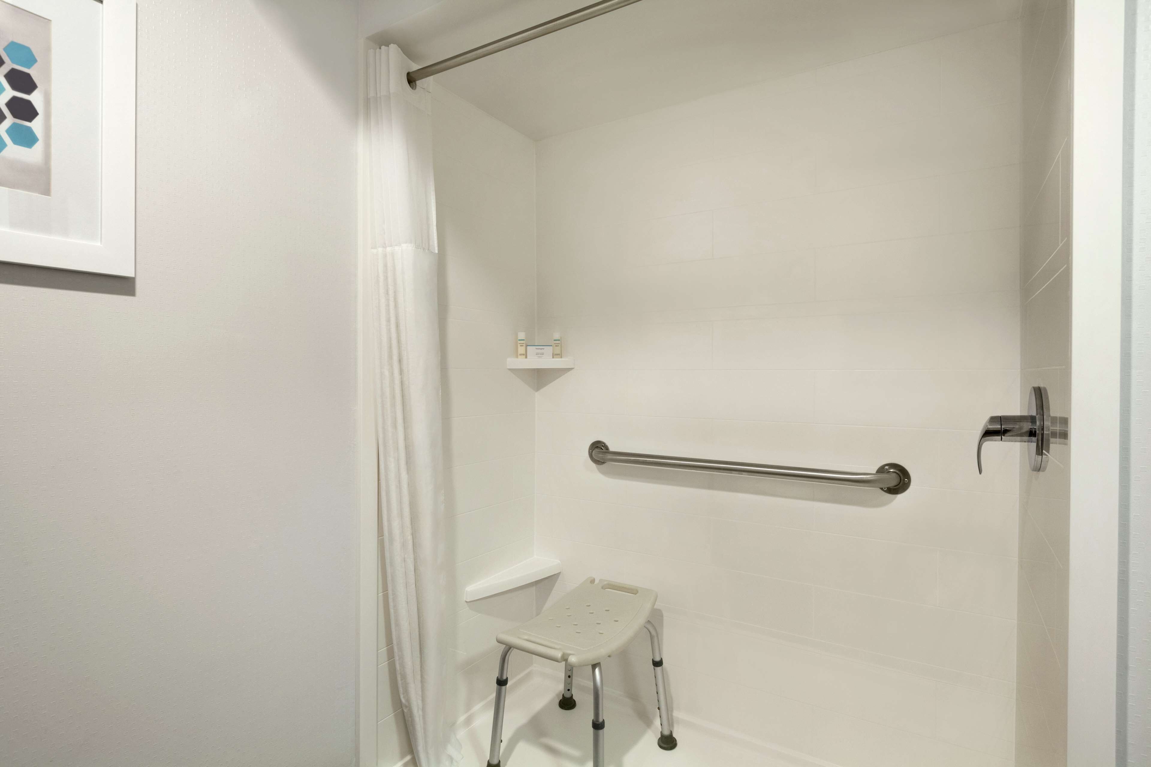 A bathroom with white walls, a shower with a glass door, a grab bar, a small white stool, and a framed picture on the wall.