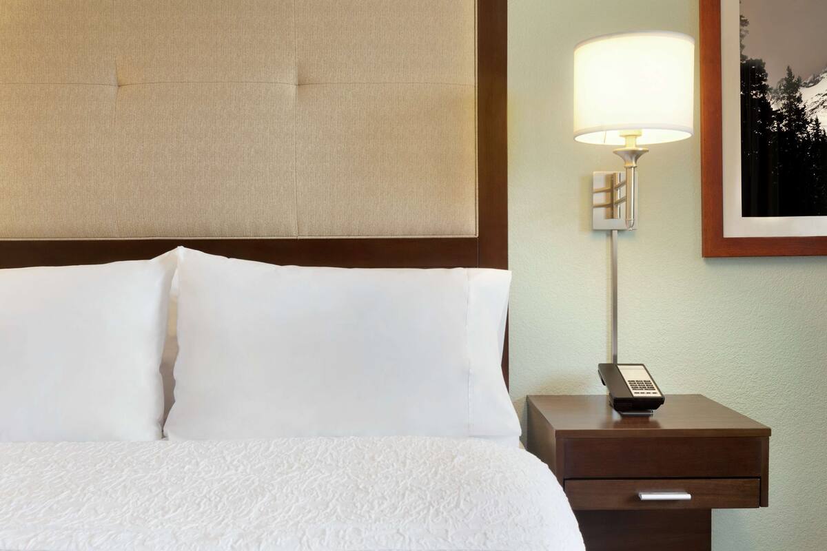 A neatly made bed with white linens, a bedside lamp, and a phone on a wooden nightstand.