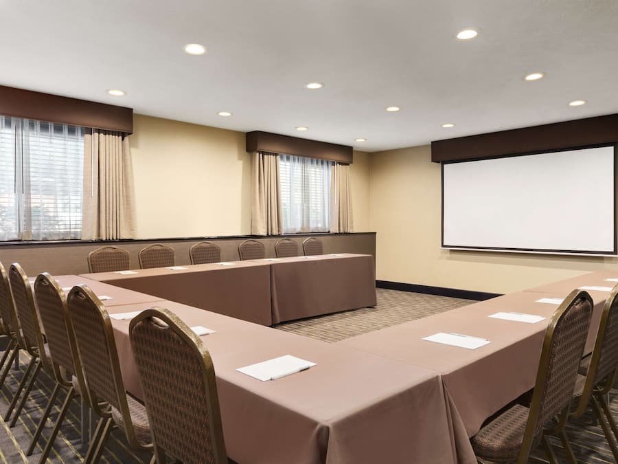 Uma sala de conferências com uma mesa comprida, cadeiras, uma tela de projetor e janelas com cortinas.