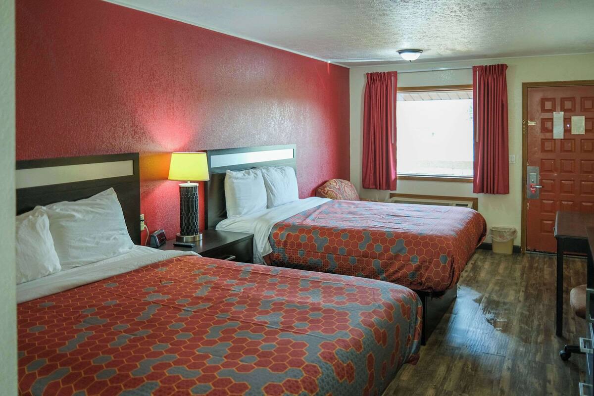A hotel room with two beds, a nightstand, a lamp, and a window with curtains.