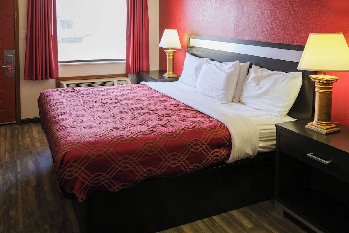 A hotel room with a large bed, two bedside tables with lamps, a wooden floor, and red curtains.