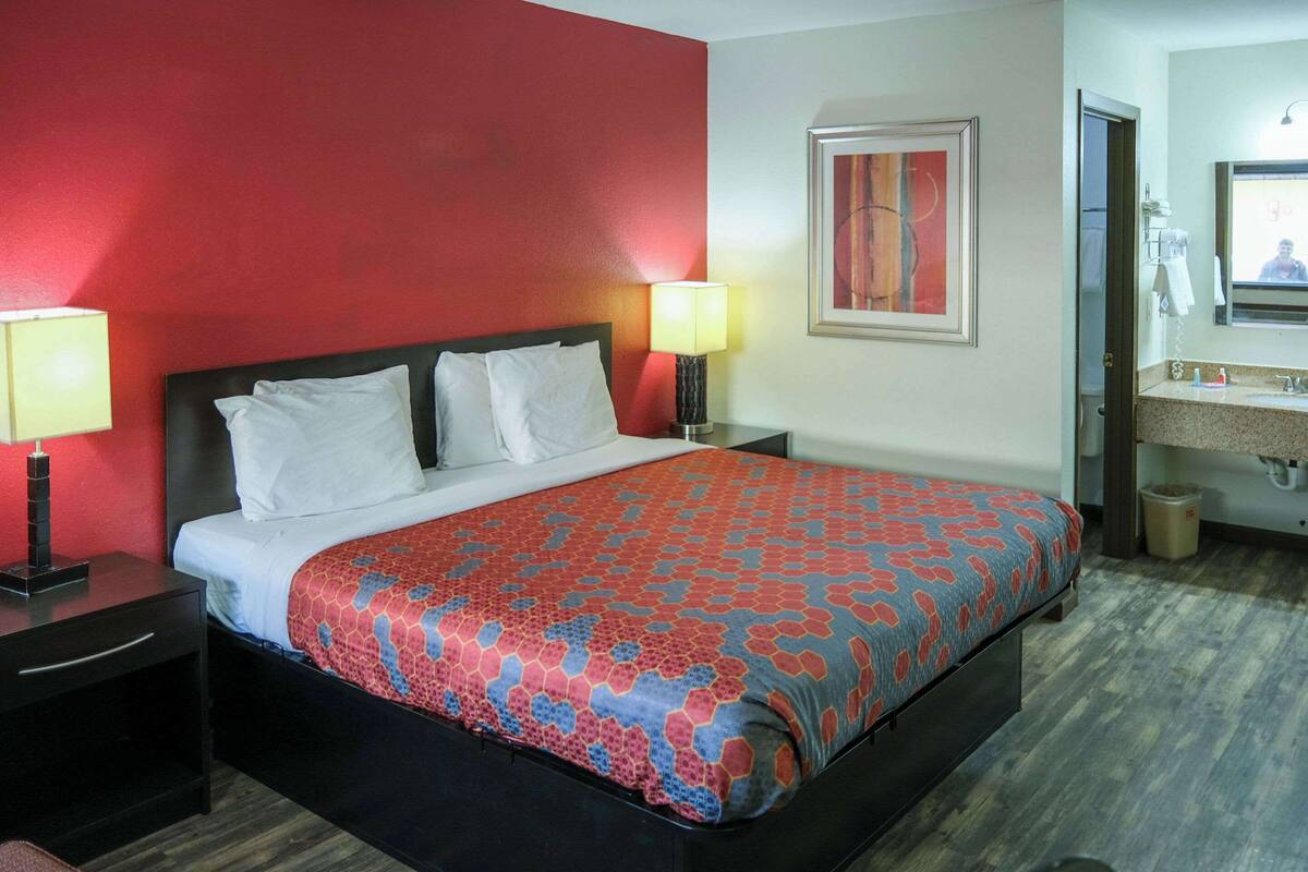 A hotel room with a bed, two bedside tables with lamps, a bathroom with a sink and mirror, and a red accent wall.