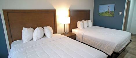 Two queen beds with white bedding, a nightstand with a lamp, and a wall-mounted picture of a bird.