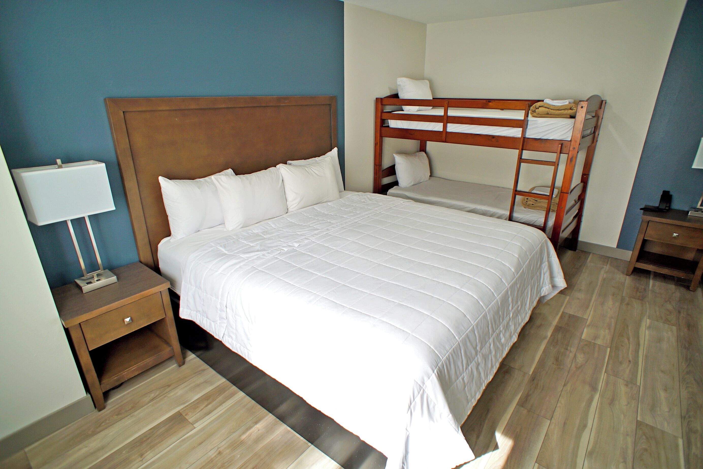 Habitación de hotel con una cama grande, una cama individual en una litera, una mesita de noche con una lámpara y piso de madera.