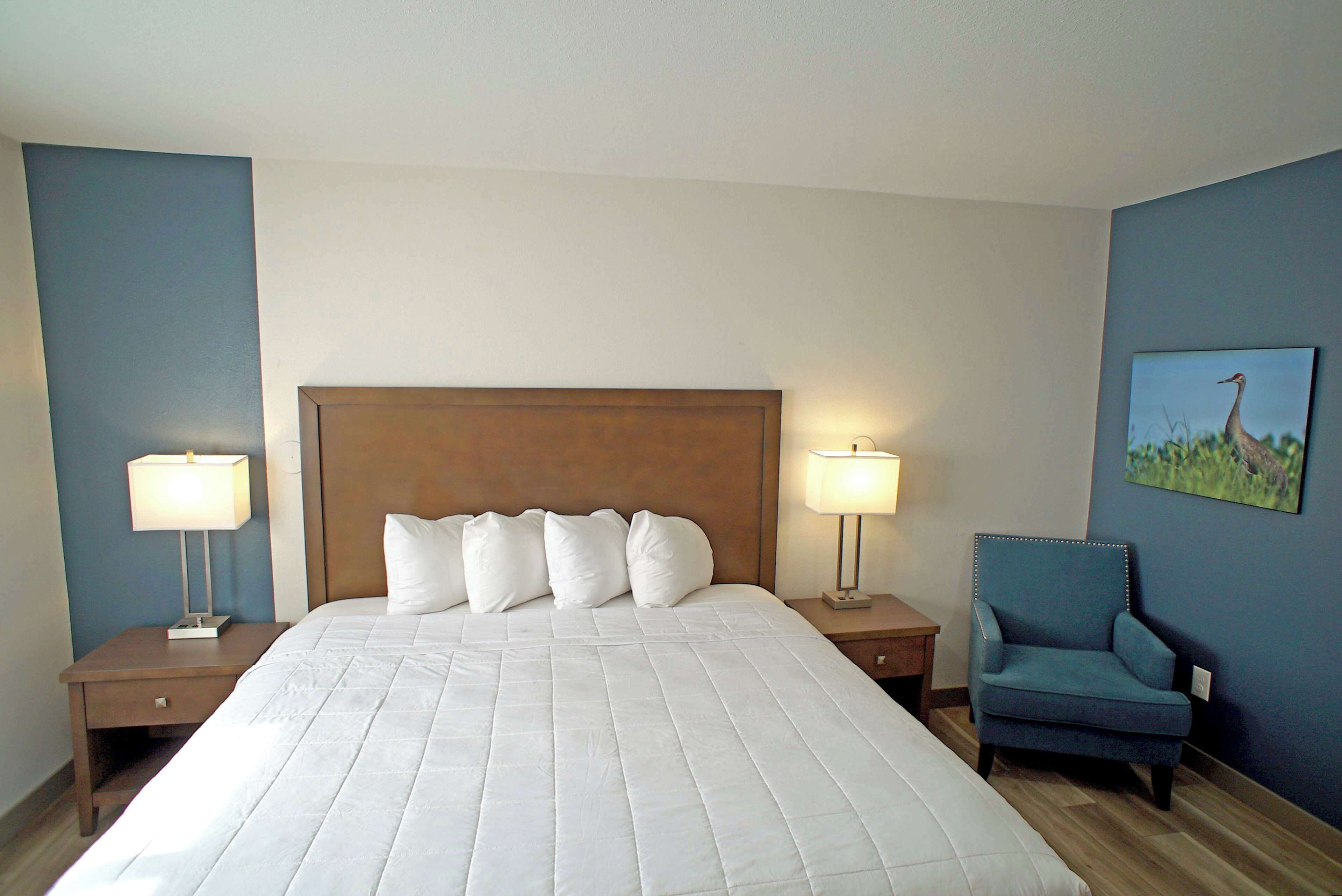 Quarto de hotel com uma cama grande, duas mesas de cabeceira com luminárias, uma cadeira e um quadro na parede.