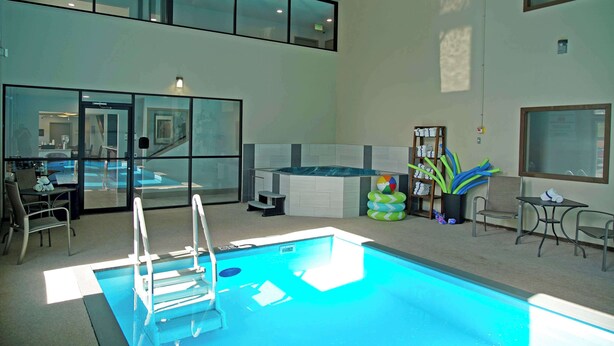 A modern indoor swimming pool with a glass wall, a hot tub, and a seating area with chairs and a table. - Baymont | Monte Vista (Monte Vista)