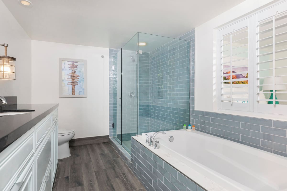 Un baño moderno con ducha de vidrio, una bañera blanca y un amplio ventanal con persianas.