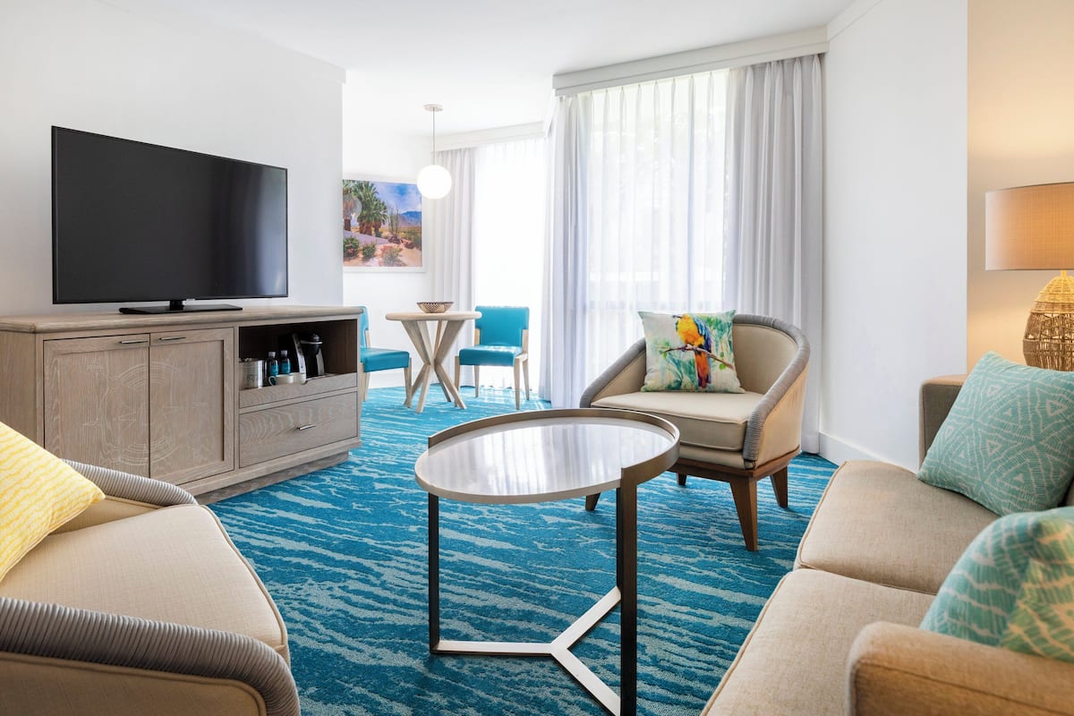 Una habitación de hotel con una televisión de pantalla plana, un mueble de madera con botellas, una alfombra azul estampada, una mesa de centro redonda, dos sillones y un sofá.