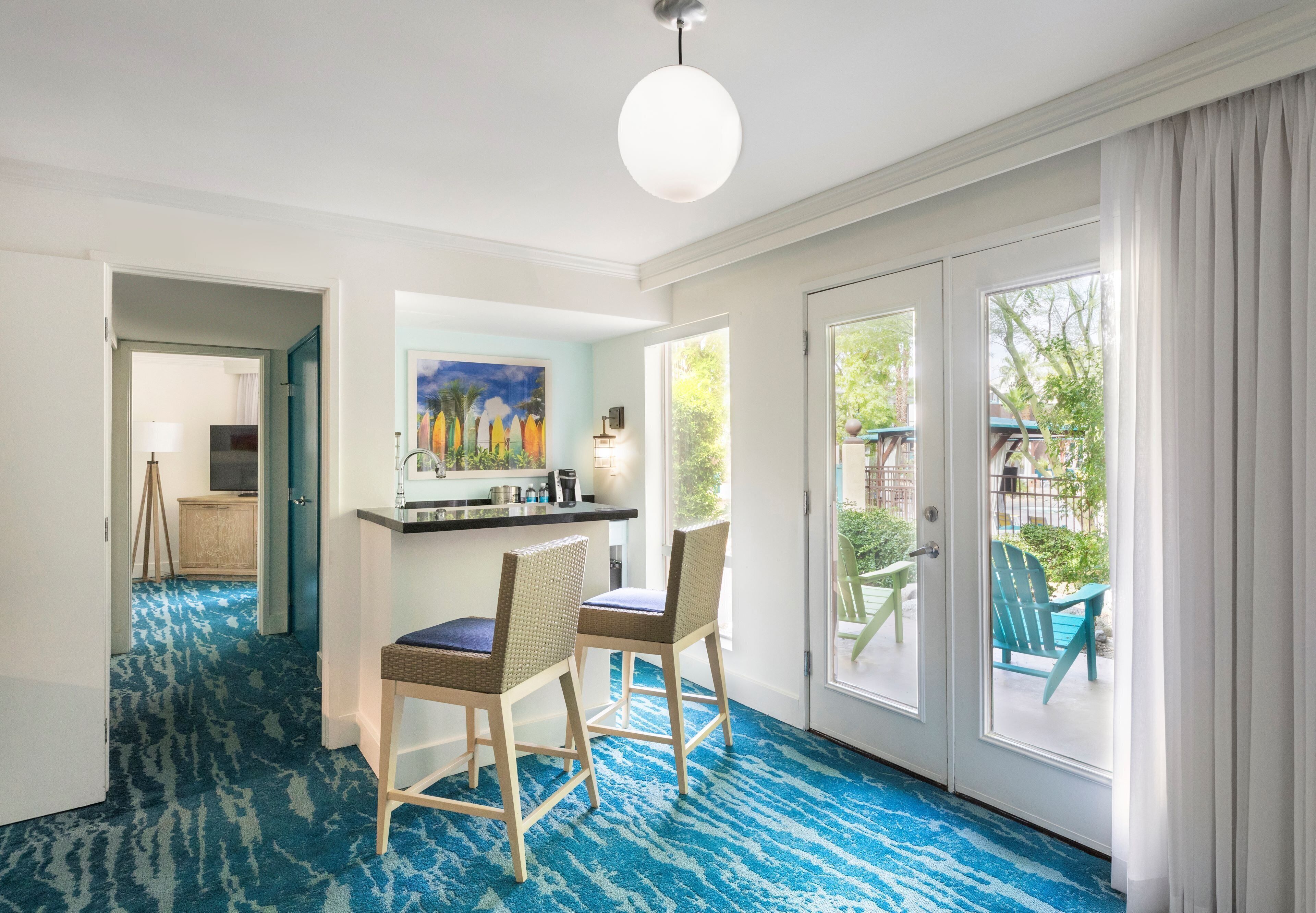 A room with a blue patterned carpet, a dining area with two chairs, a painting on the wall, and a view through a glass door.