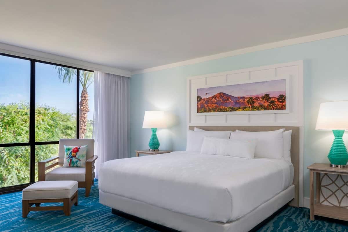Habitación de hotel con una cama grande, dos mesitas de noche con lámparas, una silla, una ventana con vistas a zonas verdes y un cuadro en la pared.