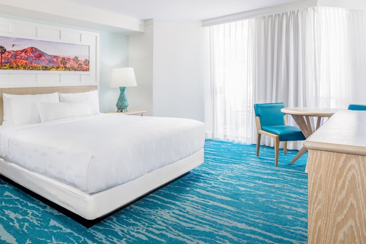 Habitación de hotel con una cama grande, un escritorio con silla, un cuadro en la pared y una ventana con cortinas.