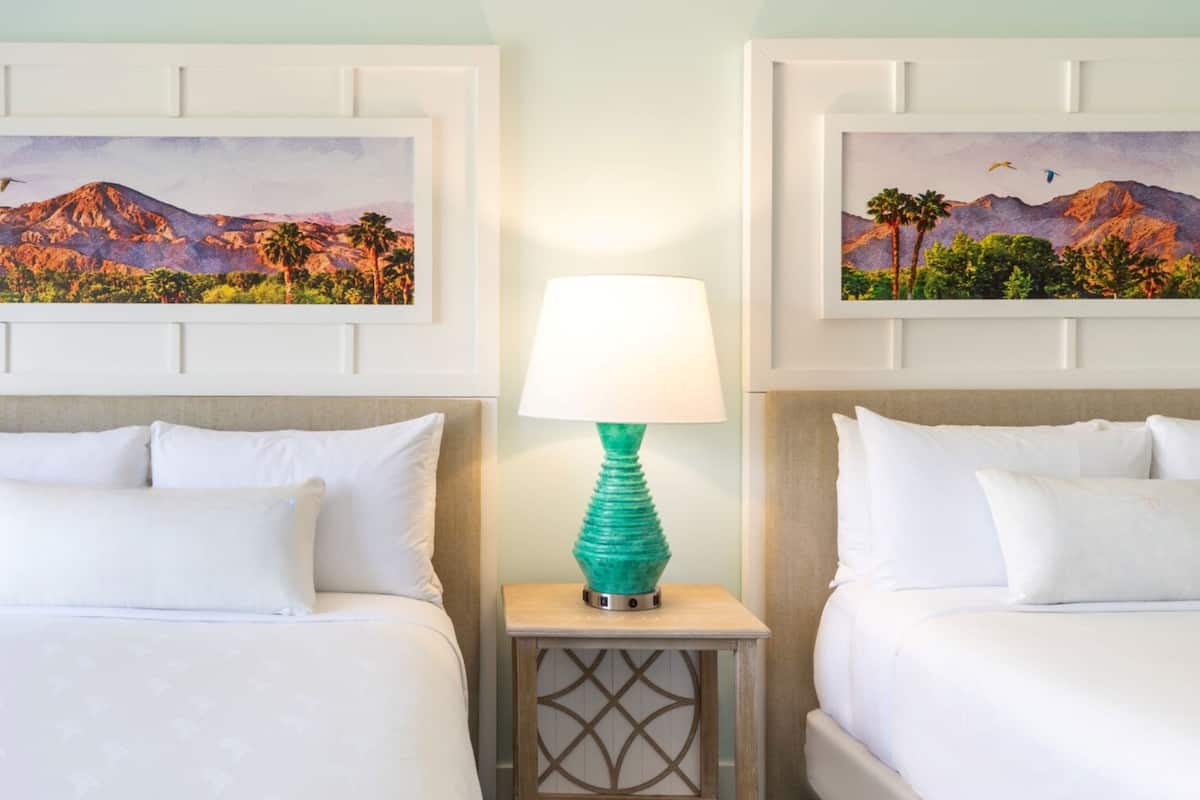 Una habitación de hotel con dos camas, una mesita de noche con lámpara y dos cuadros de paisajes enmarcados en la pared.