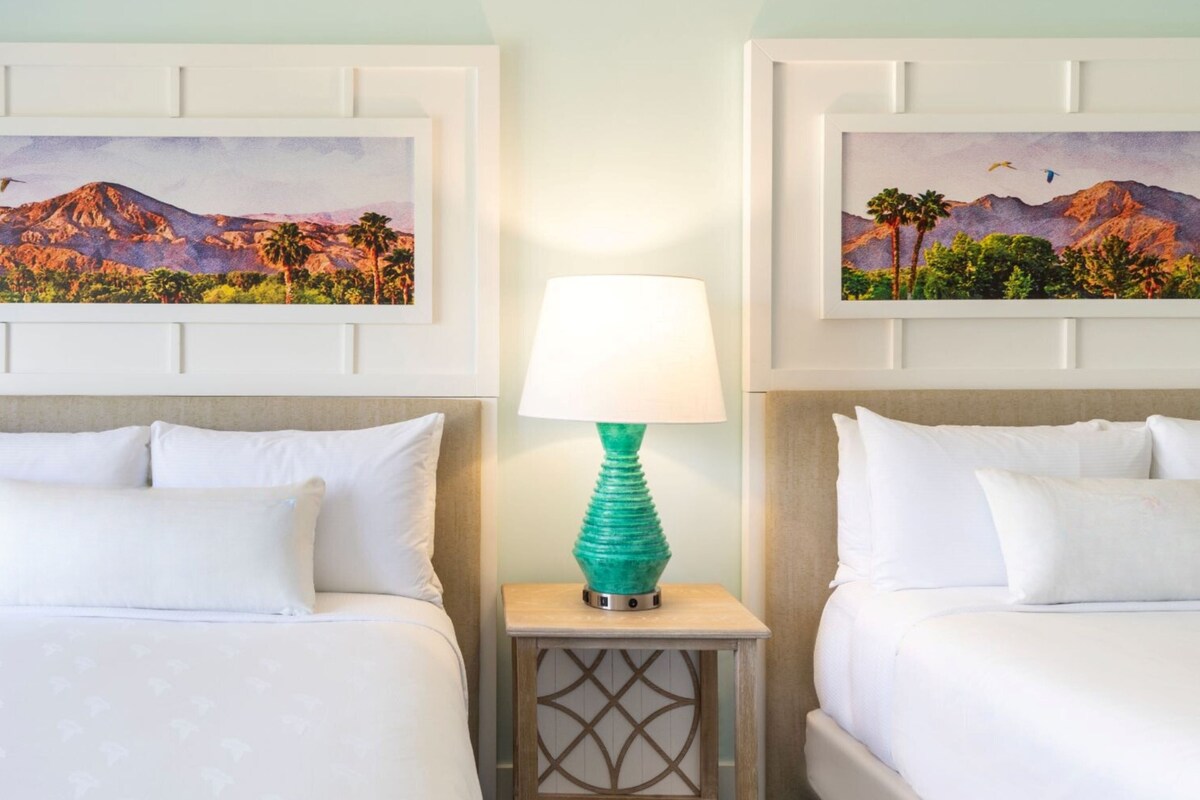 Una habitación de hotel con dos camas, una mesita de noche con lámpara y dos cuadros de paisajes enmarcados en la pared.