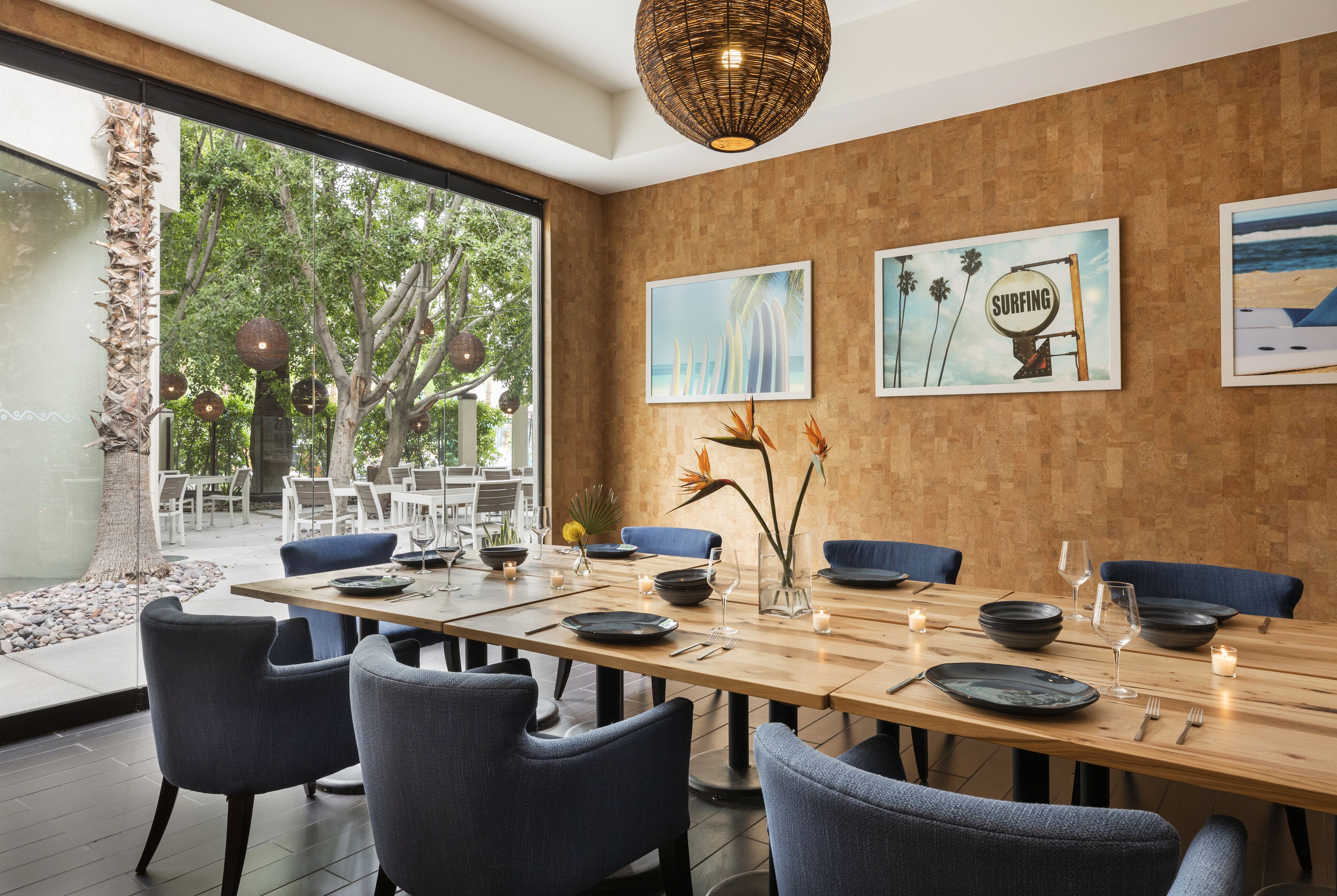 Un espace repas moderne avec une longue table dressée pour six personnes, entourée de chaises bleues. Des œuvres d’art encadrées ornent le mur, une grande fenêtre offre une vue sur les arbres et un luminaire suspendu éclaire l’ensemble.