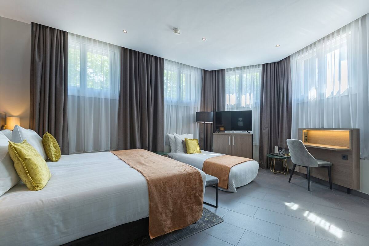 A hotel room with two beds, a desk, a chair, a TV, and large windows with curtains.