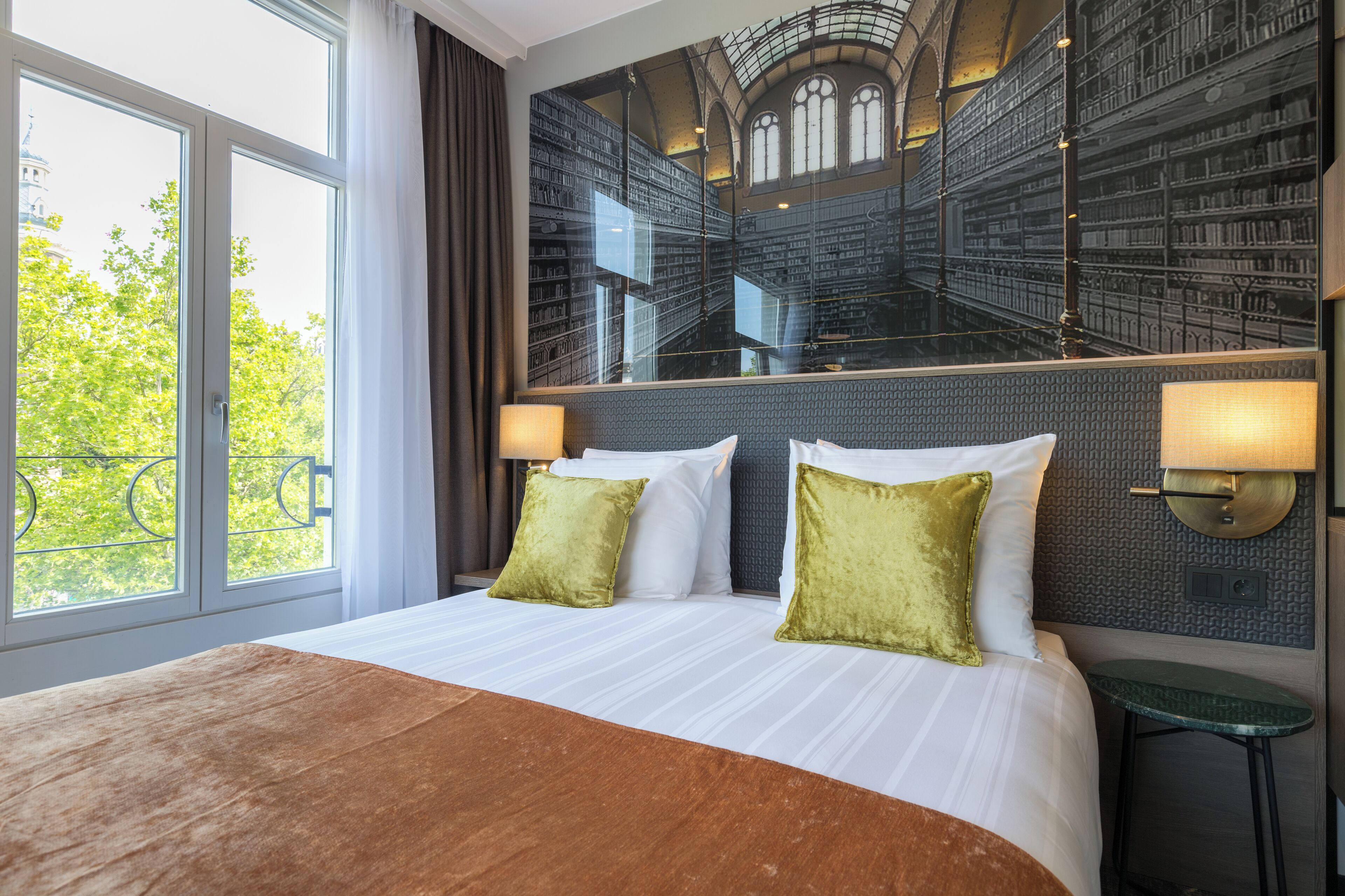 Habitación de hotel con una cama grande, dos almohadas, una mesita de noche, un espejo y vista a una biblioteca a través de la ventana.