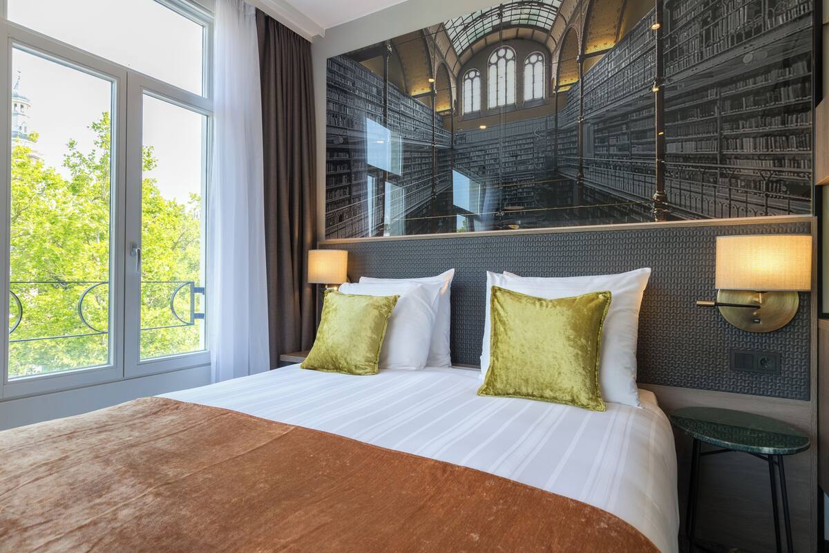 A hotel room with a large bed, two pillows, a nightstand, a mirror, and a view of a library through the window.