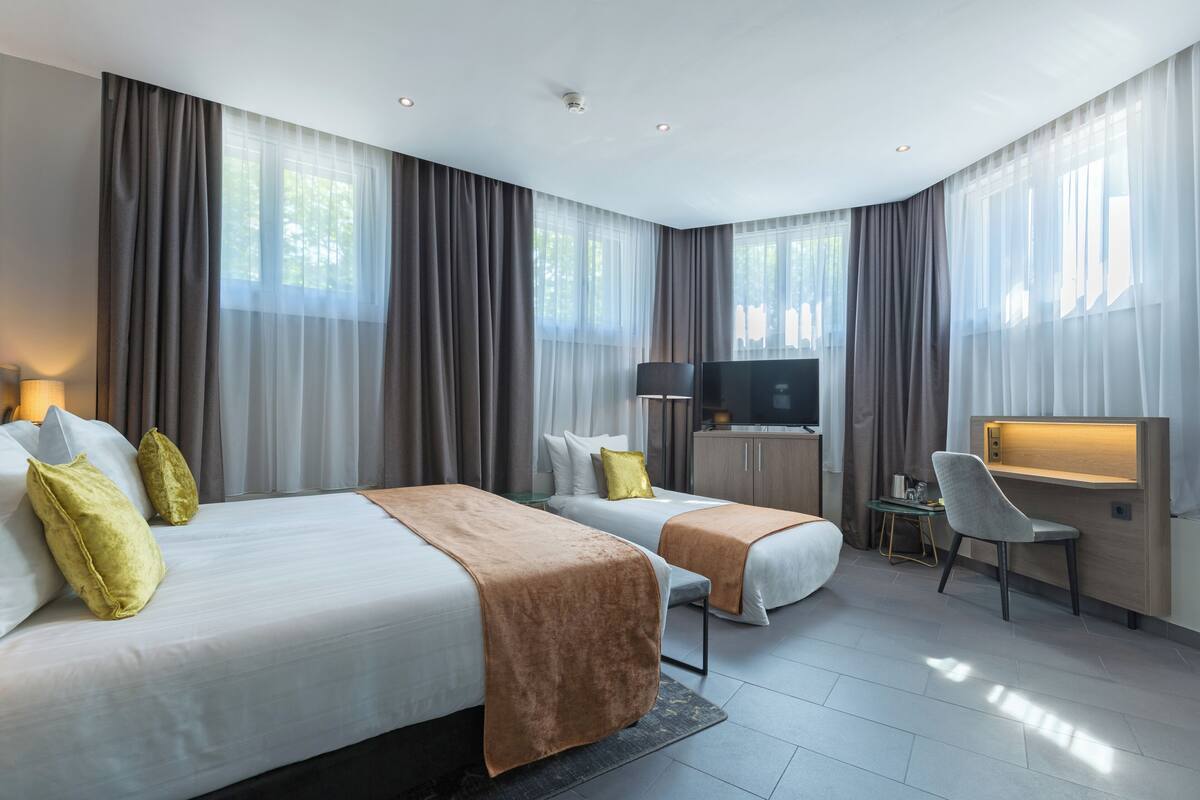 A modern hotel room with two beds, a desk, a chair, a television, and large windows with curtains.