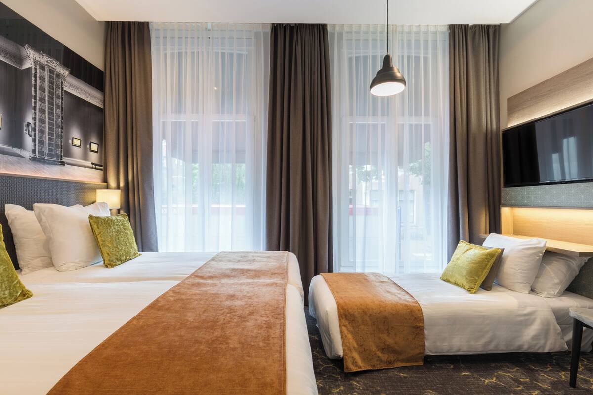 A hotel room with two beds, a large window with curtains, a flat-screen TV mounted on the wall, and a hanging pendant light.