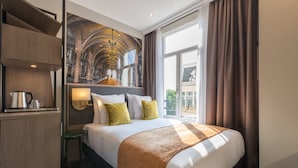 Habitación de hotel con una cama grande, un escritorio con hervidor, una mesita pequeña y un ventanal con cortinas.