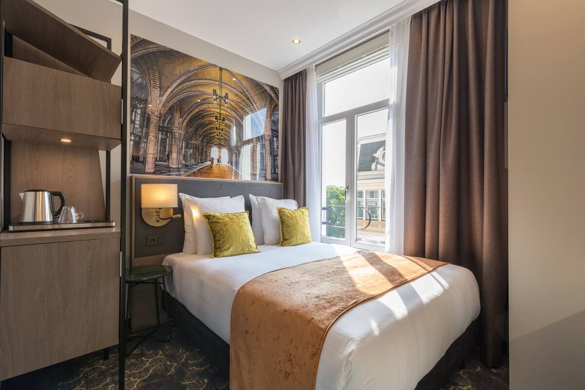 A hotel room with a large bed, a desk with a kettle, a small table, and a large window with curtains.