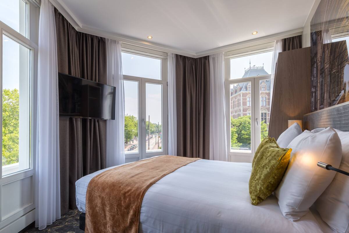 A hotel room with a large bed, a flat-screen TV mounted on the wall, and a view of the city through large windows.