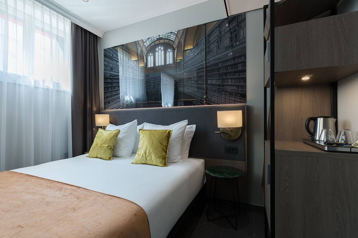 A hotel room with a large bed, two pillows, a nightstand with a lamp, a coffee maker, and a view of a library through a window.