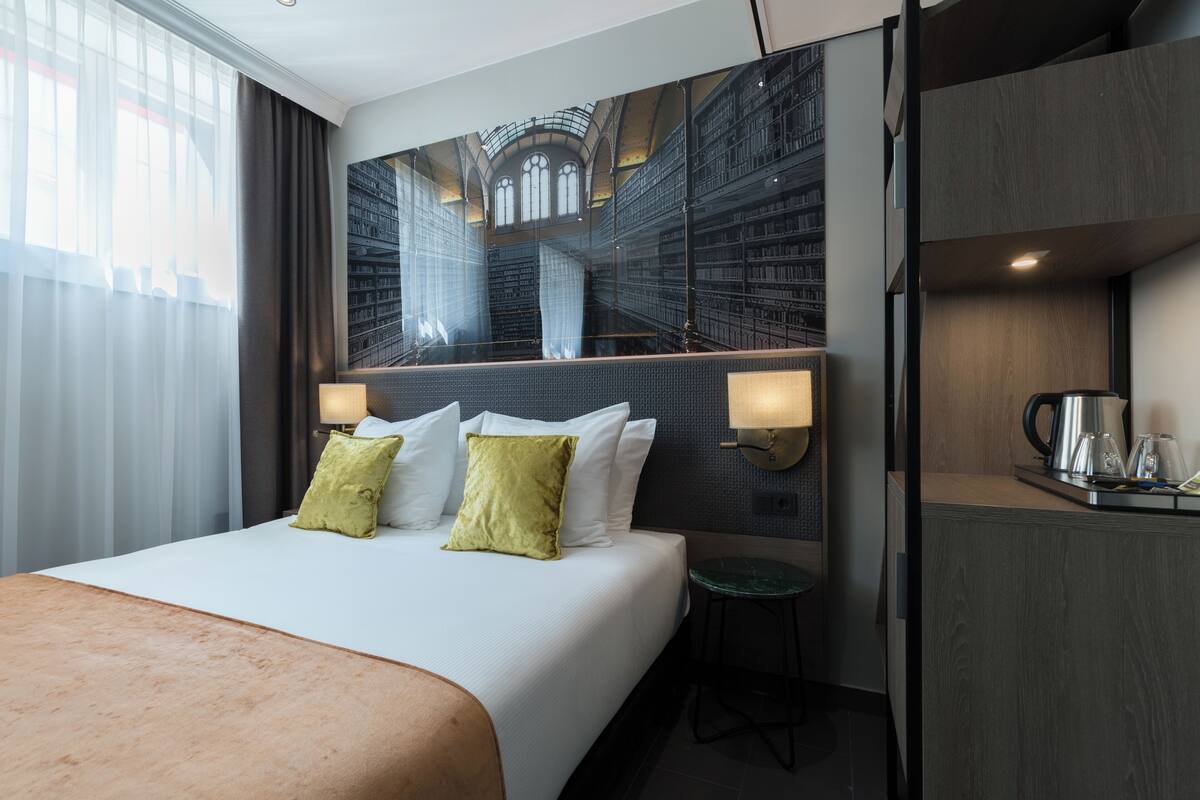 A hotel room with a large bed, a desk with a kettle, and a view of a library through a glass wall.