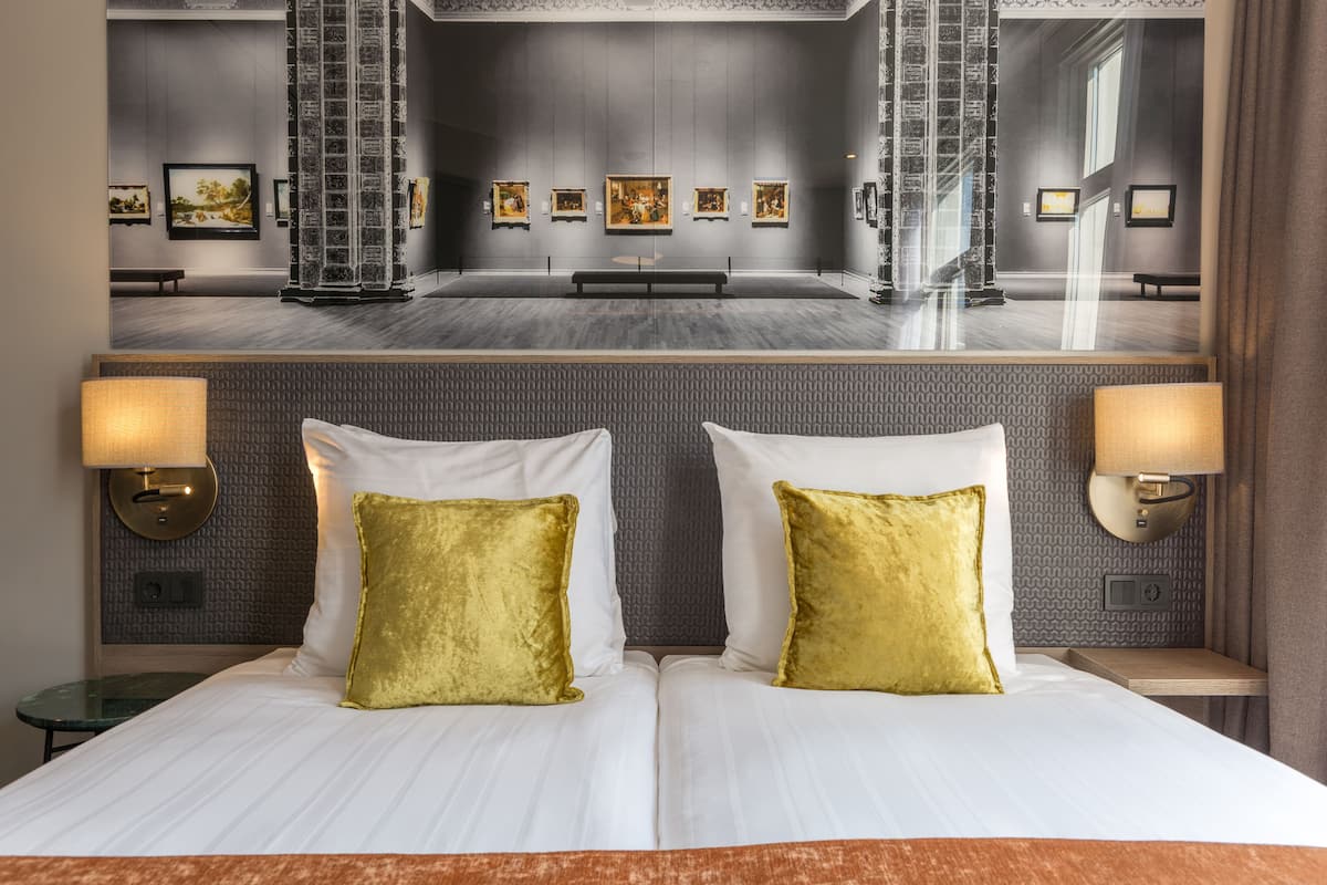 A hotel room with a large bed, two gold pillows, a nightstand with a lamp, and a wall with framed artwork.