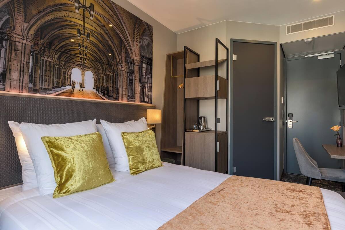 A hotel room with a large bed, a headboard with a textured design, a wall-mounted artwork depicting a historical scene, a wooden wardrobe, a desk with a chair, and a door with a handle.