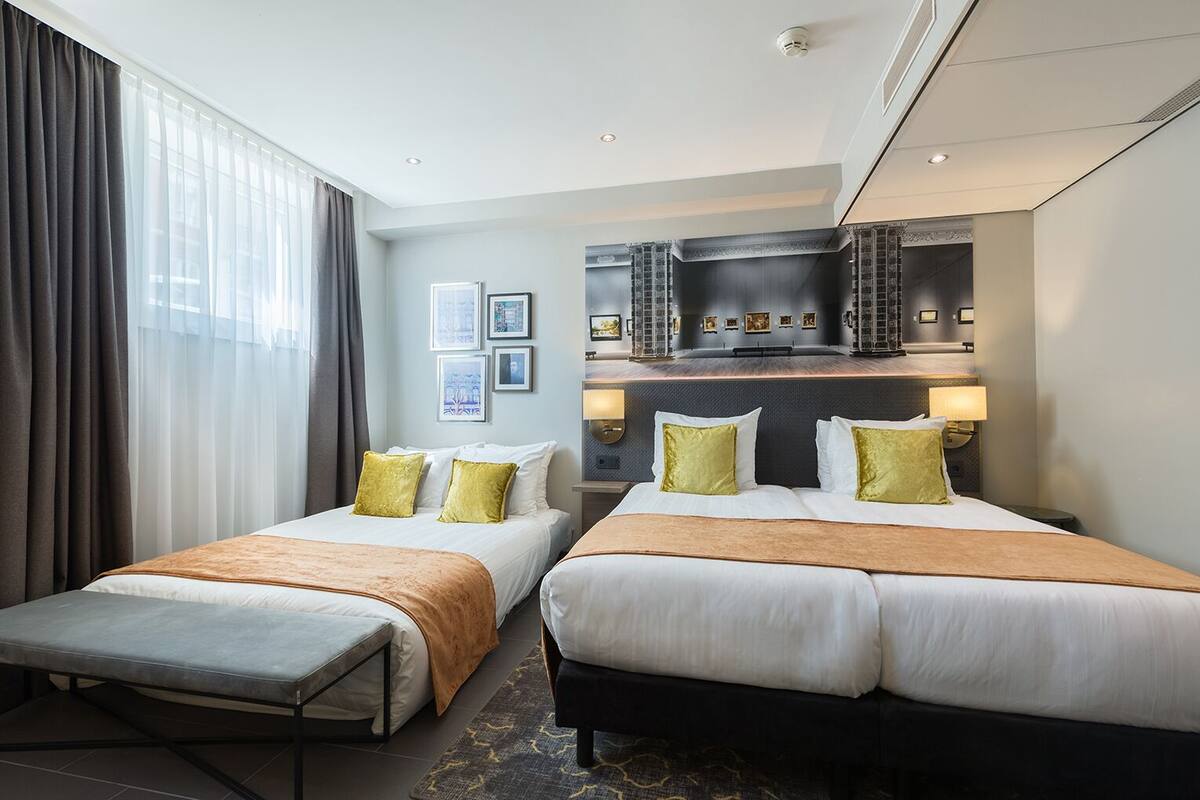 A hotel room with two beds, a bench, a minibar, and a large window with curtains.