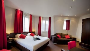 Habitación de hotel con una cama grande, un sofá, un escritorio y una silla. Se ven ventanas con cortinas rojas y una puerta.