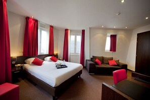 A hotel room with a large bed, a sofa, a desk, and a chair. There are windows with red curtains and a door. - Leonardo Hotel Amsterdam Museum Quarter (Amsterdam)