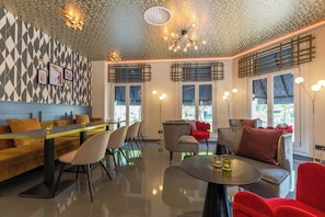 Un interior moderno con una pared de diseño geométrico, una zona de comedor con una mesa larga y sillas, un área de estar con sofás y una mesa redonda, y un techo con una lámpara original.