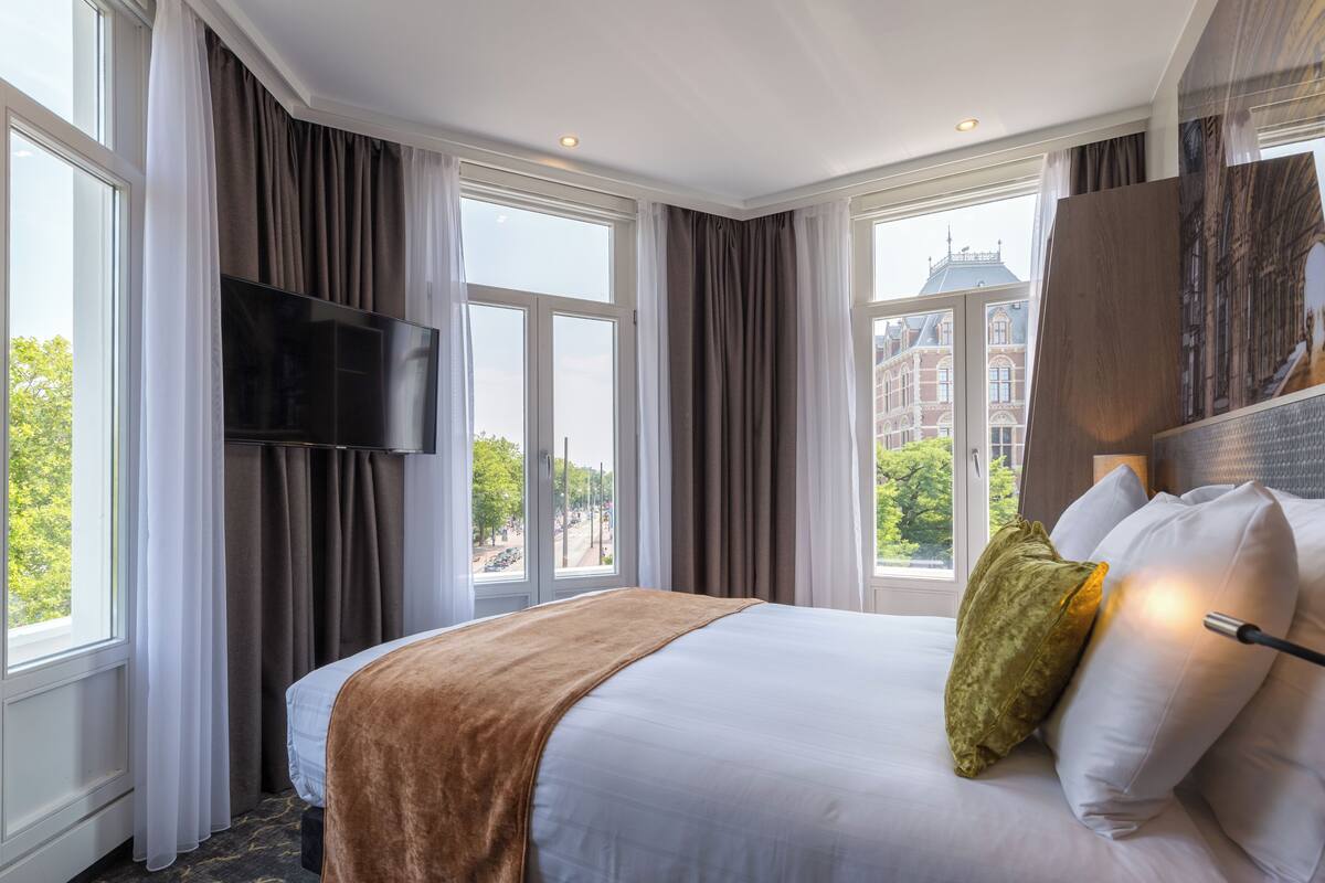A hotel room with a large bed, a flat-screen TV mounted on the wall, and a view of the city through large windows.