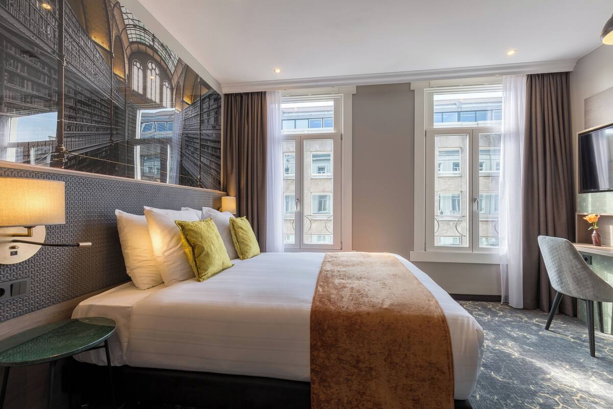 A hotel room with a large bed, a desk, a chair, a TV, and a view of the city through the windows.