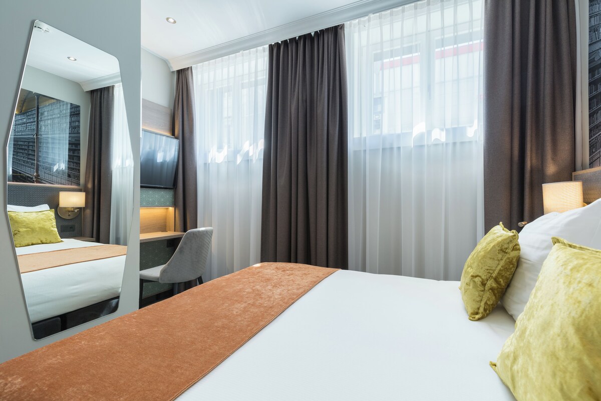 A hotel room with a bed, a desk, a chair, a large mirror, and a window with curtains.
