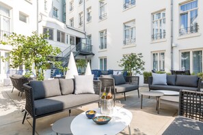 Un espace extérieur aménagé avec des canapés, des chaises et des tables, entour é de plantes en pot et bordé d’un bâtiment doté de nombreuses fenêtres et de balcons.