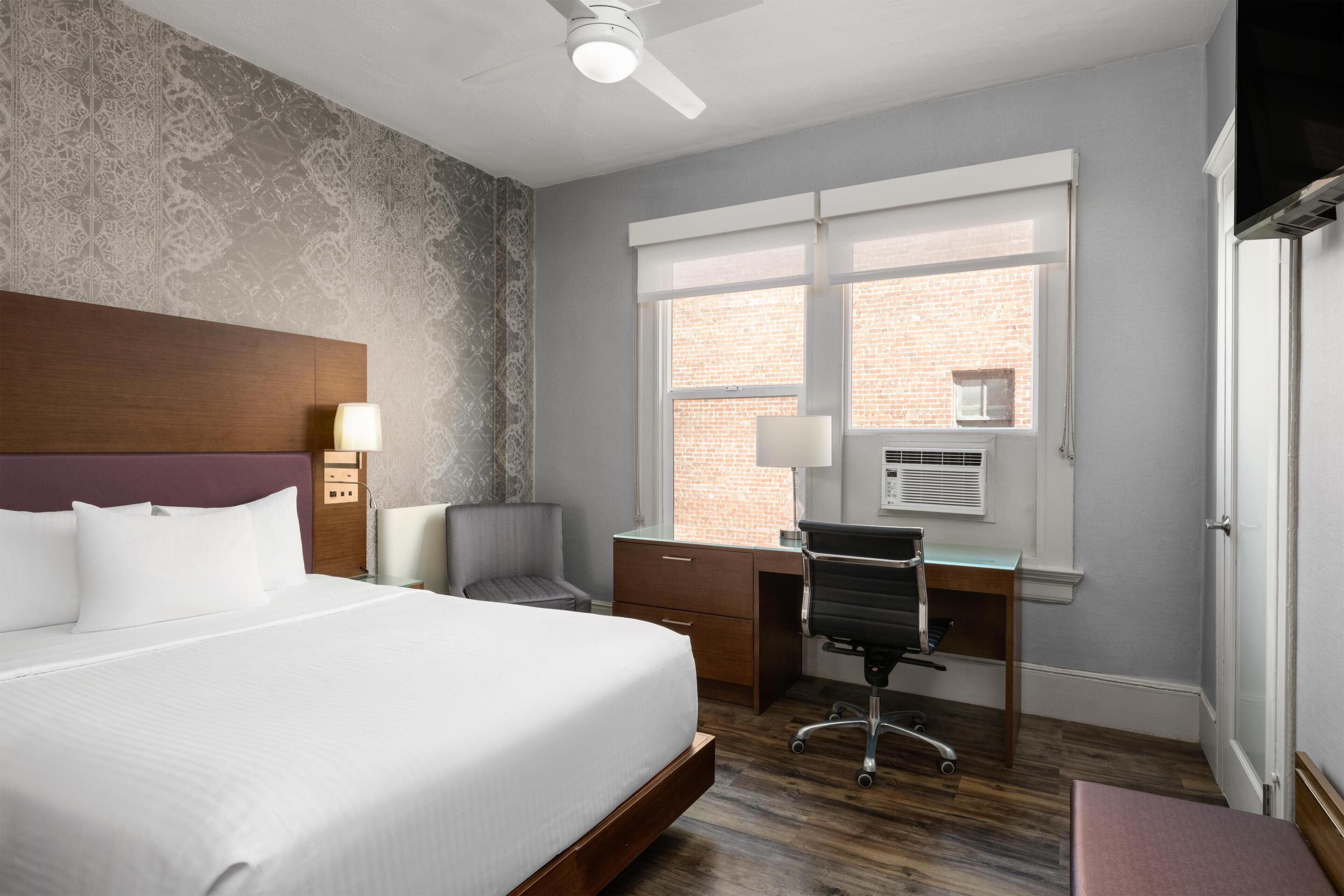 A hotel room with a bed, desk, chair, and a window with a view of a brick building.