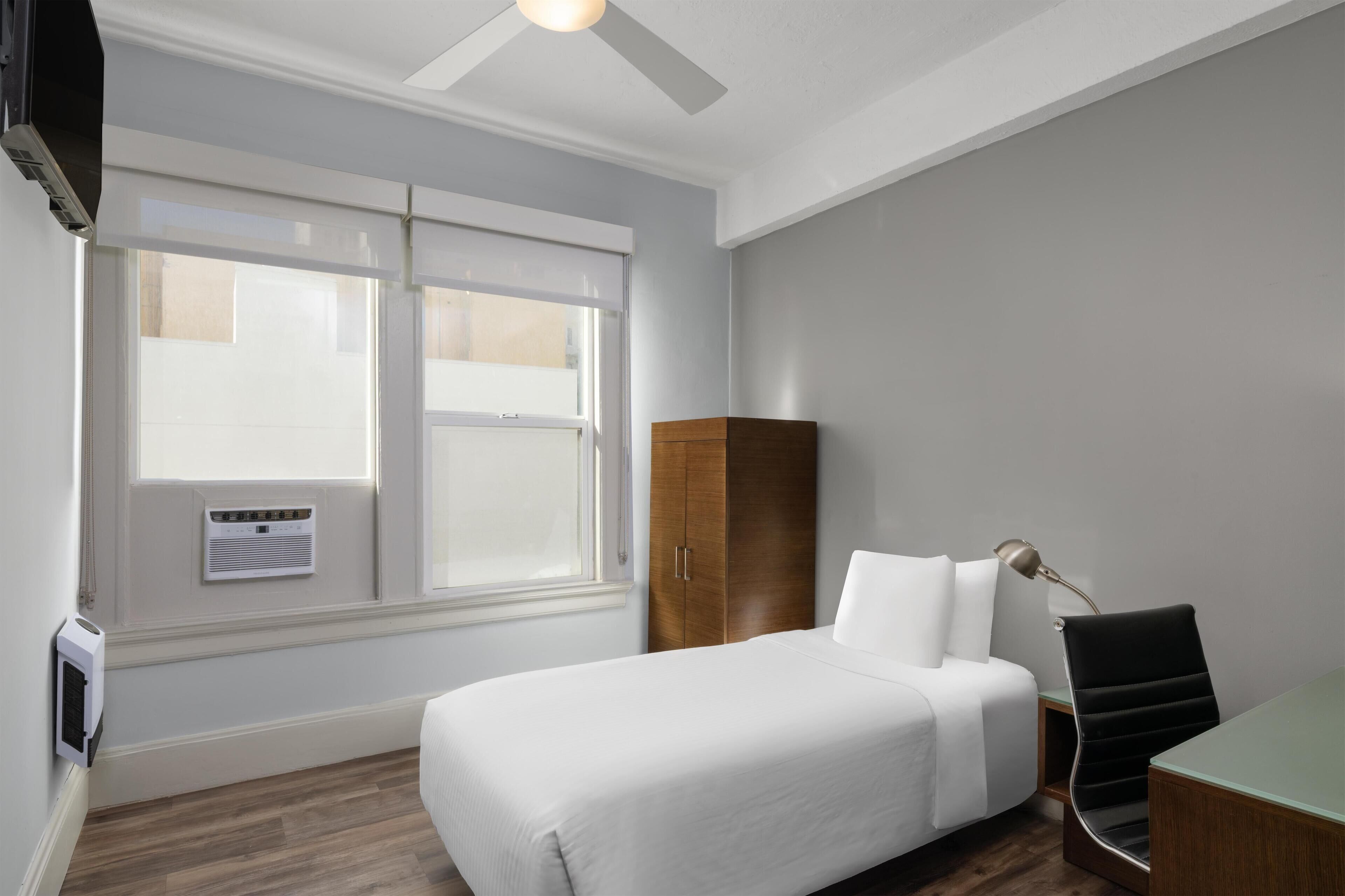 Habitació d'hotel amb un llit, una taula de treball, una cadira, un armari i un ventilador de sostre.